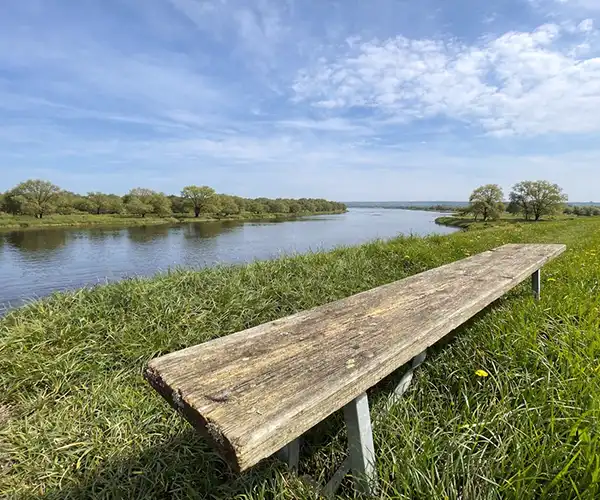 Eine Holzbank ohne Lehne an einem Fluss in einer grünen Landschaft